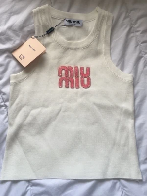 Vit linnetopp från Miu Miu - Superfin vit linnetopp från Miu Miu med broderad rosa logga framtill. Toppen är ärmlös och har en klassisk rund halsringning. Materialet är stickat och känns mjukt mot huden. Perfekt att styla med jeans eller kjol för en trendig look.