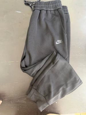 Svarta mjukisbyxor från Nike - Säljer ett par svarta mjukisbyxor från Nike med elastisk midja och dragsko. Byxorna har ribbade muddar vid bensluten, sidofickor och en diskret Nike-logga på låret. Materialet är mjukt och bekvämt, perfekt för chill dagar. Snygg och enkel design som funkar till det mesta.