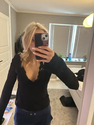 Svart långärmad topp med spets - Snygg svart långärmad topp med djup v-ringning och spetsdetaljer längs halsringningen. Tunn och figurnära modell som passar perfekt till jeans eller kjol. Enkel men ändå elegant design.