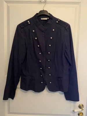 Mörkblå kavaj med silverknappar - Stilren mörkblå kavaj från Gianini med militärinspirerad design. Kavajen har ståkrage, dubbla rader av silverfärgade knappar framtill och dekorativa axelpartier. Tillverkad i ett mjukt tyg som ger en snygg passform och cool look.