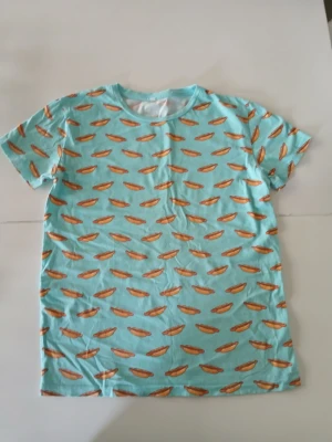 Turkos t-shirt med korvmönster - Cool turkos t-shirt med ett lekfullt mönster av korvar över hela plagget. T-shirten har rund halsringning och korta ärmar. Tillverkad i mjuk bomull som känns skön mot huden. Perfekt för dig som vill sticka ut med en unik och rolig stil.