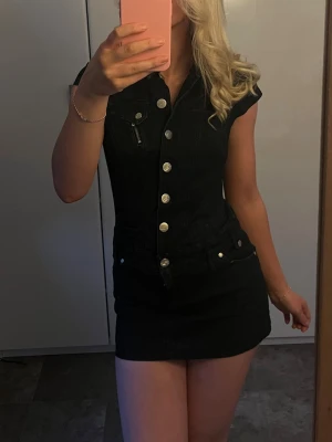 Svart jeansklänning med knappar - Snygg svart kort jeansklänning med silvriga knappar framtill och korta ärmar. Säljer då den är för kort för mig (är 175cm) köpt på secondhand. Mörkblå nästan svart 