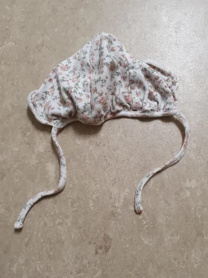 Mössa - Söt mössa från Newbie i mjuk bomull med vitt tyg och små blommor i rosa och grönt. Mössan har knytband under hakan och en klassisk form som passar perfekt till vår och sommar. Perfekt accessoar för en gullig och romantisk look.