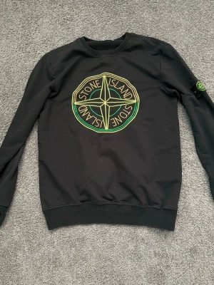 Svart Stone Island sweatshirt - Svart sweatshirt från Stone Island med stort gult och grönt logotryck på bröstet och klassisk patch på ärmen. Tröjan har rund halsringning, ribbade muddar och en avslappnad passform. Perfekt för dig som gillar streetwear och vill ha en ikonisk look.