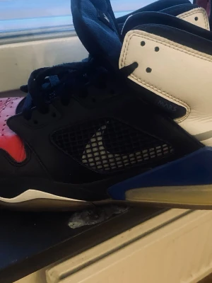 Nike Jordan Mars 270 svart/blå/röd - Nike Jordan Mars 270 sneakers med högt skaft, svart mesh och läder, blå och röda detaljer samt vit häl. Swoosh-logga på sidan och perforerad tå. Snygg och unik färgkombination som sticker ut.