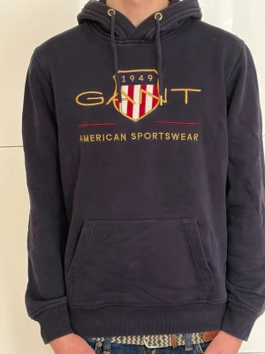 Mörkblå hoodie från GANT - Snygg mörkblå hoodie från GANT med broderad logga och texten 'American Sportswear' på bröstet. Klassisk huva med dragsko och stor magficka framtill. Tillverkad i mjukt material, perfekt för en avslappnad stil.