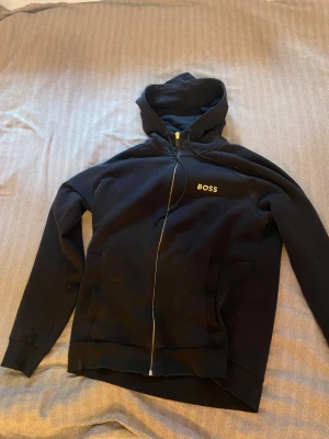 Svart hoodie med dragkedja från BOSS - Snygg svart hoodie från BOSS med dragkedja framtill och gul broderad logga på bröstet. Hoodien har huva med snörning och två fickor på sidorna. Perfekt för en chill och stilren look. Storlek M