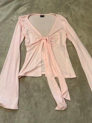 Ljusrosa omlottopp med knyt - Supersöt ljusrosa omlottopp med långa ärmar och knyt framtill. Toppen har en djup v-ringning och är gjord i ett mjukt, tunt material som känns skönt mot huden. Perfekt att styla med jeans eller kjol för en trendig look.