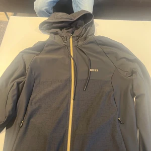 Svart hoodie från BOSS med dragkedja - Snygg svart hoodie från BOSS med hel dragkedja i kontrastfärg och diskret mönster på framsidan. Tröjan har huva med snörning och två fickor med dragkedja. Perfekt för en stilren och sportiga look