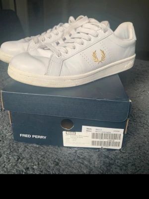 Vita Fred Perry sneakers i läder - Stilrena vita sneakers från Fred Perry i läder med guldfärgad lagerkrans på sidan och hälen. Snygg och enkel design som passar till det mesta. Skorna är sparsamt använda och i bra skick , se bilder . Skickas med originalkartongen . Nypris 1490 kr 