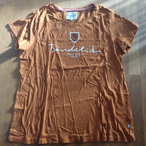 Brun t-shirt från Bondelid - Snygg brun t-shirt från Bondelid med vitt tryck och logga på bröstet. Klassisk rund halsringning och korta ärmar. Tillverkad i mjuk bomull som känns skön mot huden. Perfekt för en avslappnad och stilren look.