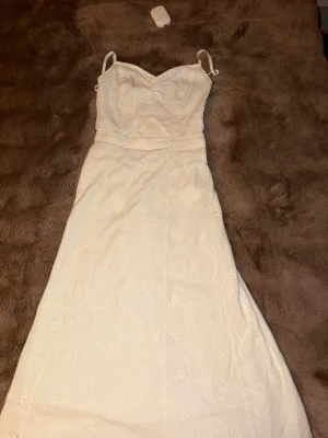 Vit långklänning från Hollister - Supersöt vit långklänning från Hollister med smala axelband och broderade detaljer. Klänningen har en feminin passform och är perfekt för varma dagar. Tillverkad i bomull för en skön och luftig känsla.