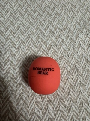 Romantic Bear orange läppbalsam - Säljer ett orange läppbalsam från Romantic Bear i rund förpackning. Balsamet har en smidig klotformad design som gör det enkelt att applicera. Perfekt att ha i väskan för återfuktade läppar hela dagen.