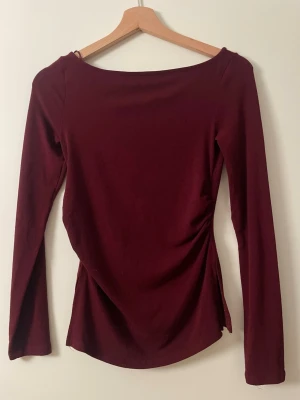 Berhska Vinröd långärmad topp med rynk - Snygg vinröd långärmad topp med rynkdetalj i sidan. Toppen har en rund halsringning och är figurnära i modellen. Perfekt att styla med jeans eller kjol för en trendig look.