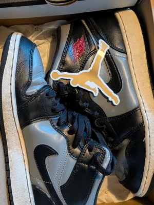 Air Jordan 1 Mid svart/grå - Snygga Air Jordan 1 Mid sneakers i svart och grått läder med klassisk röd Air Jordan-logga på sidan och svart swoosh. Skorna har svart snörning, vit mellansula och svart yttersula. Perfekta för dig som gillar streetwear och ikoniska sneakers.