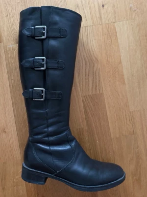 Ecco Svarta knähöga läderstövlar med spännen - Snygga svarta knähöga stövlar i äkta läder med tre dekorativa spännen på utsidan. Klassisk rund tå och låg klack. Dragkedja på insidan för enkel på- och avtagning. Perfekta för en stilren och edgy look.