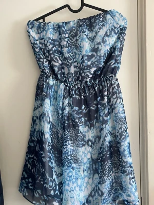 Blå axelbandslös kortklänning - Säljer en snygg axelbandslös kortklänning med blått och vitt mönster. Klänningen har resår i midjan för en bekväm passform och ett lätt, luftigt tyg som passar perfekt till varma dagar. Unik print som ger en cool vibe.