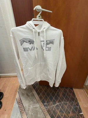 Vit hoodie med tryck från BVALDI - Vit zip-hoodie från BVALDI med svart tryck av två händer och text på bröstet. Tröjan har huva med dragsko, fickor framtill och ribbade muddar. Perfekt för en avslappnad och trendig stil. Helt ny oanvänd 