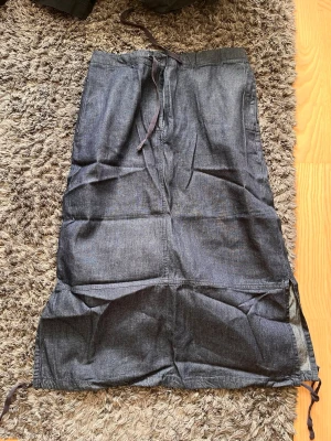 Mörkblå långkjol med snörning - Snygg mörkblå långkjol i denim-look med snörning i midjan och vid nederkanten. Kjolen har en rak passform och är perfekt för dig som gillar en avslappnad men ändå stilren vibe. Passar till både sneakers och boots.