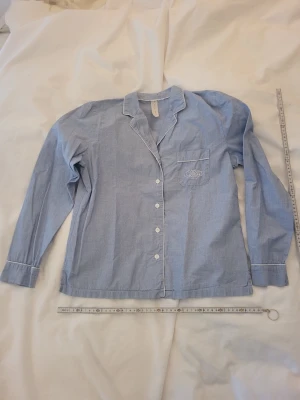 Blå långärmad pyjamaströja - Klassisk blå långärmad pyjamaströja med vita knappar och kontrastsömmar. Tröjan har en bröstficka med broderad detalj och är tillverkad i mjuk bomull. Perfekt för mysiga kvällar hemma.