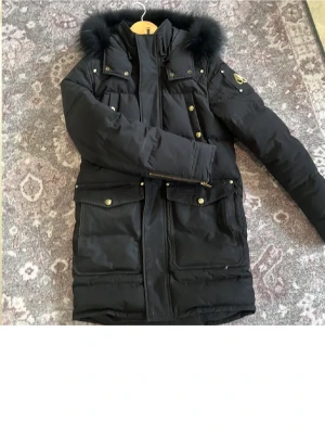 Moose Knuckles jacka - Snygg svart parkas från Moose Knuckles med avtagbar pälskant på huvan och guldfärgade detaljer. Jackan har flera stora fickor framtill, dragkedja och tryckknappar. Perfekt för kalla dagar och riktigt varm. Klassisk logga på ärmen.