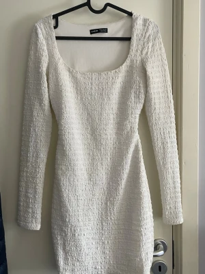 Vit långärmad klänning - Trendig vit klänning med lång ärm och strukturerat tyg. Klänningen har en fyrkantig ringning fram och öppen ryggdetalj. Kort modell som sitter snyggt på kroppen och passar perfekt till vår och sommar.