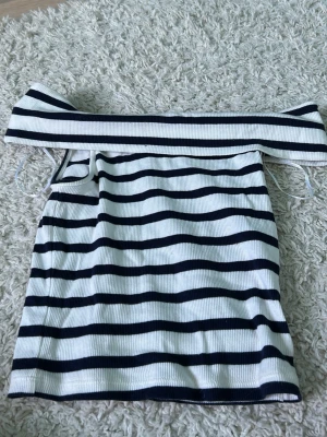 Randig offshoulder topp i ribbad bomull - Säljer en vit och svart ribbad offshoulder topp med horisontella ränder. Toppen har snörning i sidorna och är axelbandslös för en trendig look. Perfekt för dig som gillar en stilren och modern vibe.
