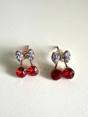 Söta körsbärsörhängen med strass, helt NYA - Ett par örhängen i form av körsbär med glittrande röda pärlor som bär och en liten rosett upptill dekorerad med skimrande strasstenar. Guldfärgad metall ger en lyxig touch och örhängena har stift. Perfekta för dig som gillar lekfulla accessoarer. Helt NYA oanvända. 