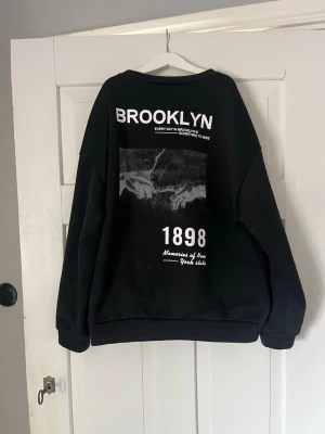 Svart sweatshirt med Brooklyn-tryck - Cool svart sweatshirt med stort Brooklyn-tryck och grafiskt motiv på ryggen. Texten '1898' och 'Memories of New York side' ger en urban vibe. Tröjan har rund hals och långa ärmar, perfekt för en avslappnad streetwear-look.
