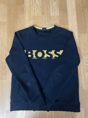 Svart sweatshirt från BOSS - Stilren svart sweatshirt från BOSS med gul logga framtill. Tröjan har rund halsringning, långa ärmar och ribbade muddar vid ärmslut och nederkant. Perfekt för en avslappnad och trendig look.