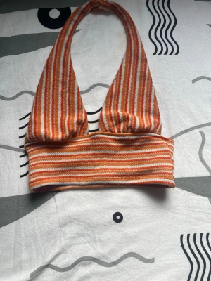 Randig ribbad halterneck topp - Säljer en ribbad halterneck topp från H&M med smala ränder i orange, vitt och beige. Toppen är ärmlös och har en croppad passform, perfekt för varma dagar. Materialet är mjukt och stretchigt för en bekväm känsla.