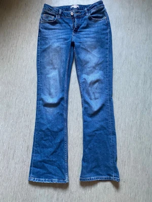 Blå bootcut jeans med fickor - Snygga blå bootcut jeans med klassiska fem fickor och knappgylf. Jeansen har en lätt tvättad look och raka ben som går ut lite nedtill. Perfekta för en avslappnad och trendig stil. Materialet är denim i bomull.