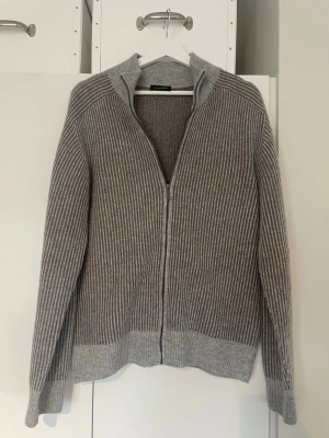 Grå ribbstickad kofta Sand - Snygg grå ribbstickad kofta från Sand med dragkedja framtill. Koftan har hög krage, långa ärmar och breda muddar i ljusare grått. Perfekt för lager på lager och enkel att matcha med olika outfits.                                                                            Kontakta gärna vid fler frågor eller bilder!