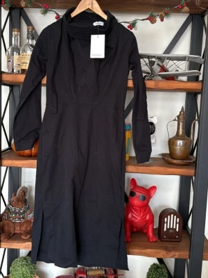 Svart långärmad klänning Rodebjer - Stilren svart långklänning från Rodebjer med långa ärmar och hög krage med knytband. Klänningen har slitsar nedtill på båda sidor och en rak, elegant siluett. Perfekt för dig som gillar minimalistisk design och tidlösa plagg. Helt ny. 