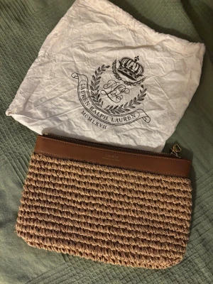 Flätad kuvertväska från Lauren Ralph Lauren - Snygg kuvertväska från Lauren Ralph Lauren i naturfärgat flätat material med bruna läderdetaljer och guldfärgad dragkedja. Perfekt storlek för det viktigaste och ger en trendig touch till din outfit. Kommer med original dustbag.