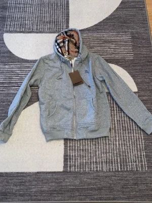Grå hoodie med Burberry-mönster - Säljer en grå hoodie från Burberry med dragkedja framtill och klassiskt rutigt foder i huvan och insidan. Hoodien har fickor på sidorna, ribbade muddar och justerbar huva med snören. Perfekt för dig som vill ha en stilren och lyxig look.