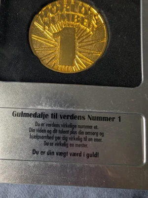 Guldmedalj World's Number 1 - Säljer en guldmedalj med texten 'World's Number 1' i relief. Medaljen hänger i ett rött och vitt band och levereras i en silverfärgad presentförpackning med dansk text. Perfekt som rolig present till någon du vill hylla.