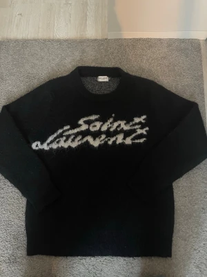 Svart Saint Lauren stickad tröja - Säljer en svart stickad tröja från Saint Lauren med vit logotyp i borstigt garn framtill. Tröjan har rund halsringning och långa ärmar. Materialet ser ut att vara mjukt och fluffigt, perfekt för kyligare dagar.
