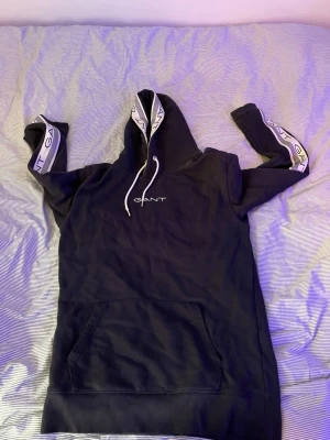 Svart hoodie från GANT med logga - Svart hoodie från GANT med vit logga på bröstet och vita detaljer längs huvan och ärmarna där det står GANT. Klassisk känguruficka framtill och vita dragsnören i huvan. Skön och stilren modell som passar perfekt till jeans eller mjukis.