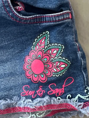 Blå jeansshorts Sun & Sand blommig - Snygga blå jeansshorts med rosa och turkosa detaljer, blommigt tryck och broderi. Shortsen har fransig kant, rosa sömmar och spetsdetaljer samt knappstängning. Perfekta för varma dagar! 