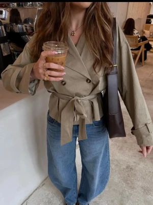 Kort trenchcoat - Snygg kort trenchcoat i beige med dubbelknäppning, bred krage och knytskärp i midjan. Perfekt inför våren!