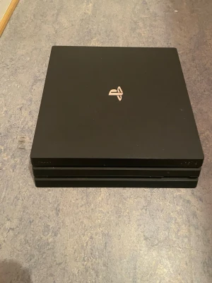 PlayStation 4 Pro med två kontroller - Säljer en svart PlayStation 4 Pro från Sony med två matchande svarta DualShock 4-kontroller. Konsolen har en stilren, fyrkantig design med PlayStation-loggan på ovansidan och är tillverkad i matt plast.