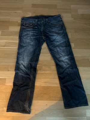 Blå jeans från True Religion - Säljer ett par blå jeans från True Religion med snygga slitningar och kontrastsömmar. Klassisk femficksmodell med raka ben och detaljerade bakfickor med lock och knapp. Jeansen har en cool tvätt och är tillverkade i denim.