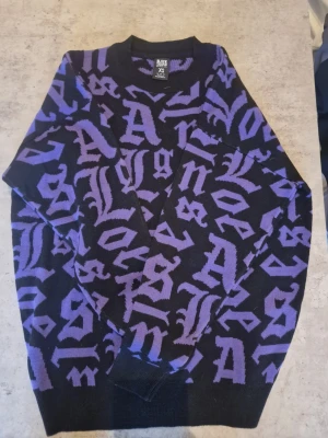 Svart och lila stickad tröja Black Squad - Xs men passar säkert medium, Stickad tröja från Black Squad i relaxed fit, svart bas med lila grafiskt bokstavsmönster över hela plagget. Rund halsringning och ribbade muddar vid ärmslut och nederkant. Perfekt för dig som gillar streetwear och unika prints.