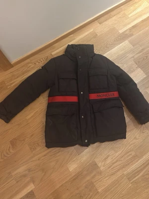 Svart Moncler dunjacka med röd detalj - Snygg svart dunjacka från Moncler med röda detaljer och logga på bröstet. Jackan har fyra stora fickor framtill, hög krage och röd insida. Perfekt för kalla dagar när du vill hålla stilen och värmen.