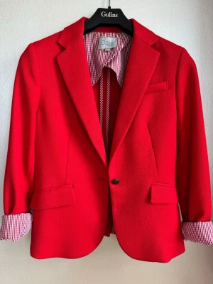 Röd kavaj med rutigt uppvik - Stilren röd kavaj från Dalia med klassisk krage, enkel knappstängningr och två fickor framtill. Kavajen har ett snyggt rödvitrutigt innerfoder som syns om man viker upp ärmsluten -  cool kontrast. Figursydd modell.