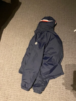 Marinblå vindjacka från Moncler - Snygg marinblå vindjacka från Moncler med huva och ikonisk logga på bröstet. Jackan har elastiska muddar, dragkedja och en sportig look med rödvit detalj på huvan. Perfekt för blåsiga dagar och enkel att matcha till olika outfits. Har ett litet hål i armen men går att sy 