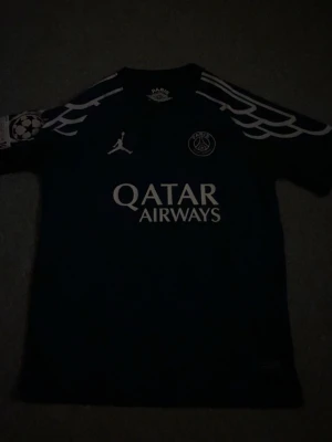 PSG matchtröja Dembele #10 - Snygg mörkblå PSG matchtröja med vita detaljer, Qatar Airways-tryck på bröstet och Champions League-märke på ärmen. Jordan-logga och PSG-emblem framtill, Dembele och nummer 10 på ryggen. Perfekt för fotbollsfans!