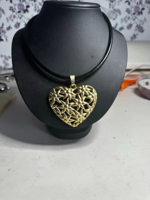 Halsband med hjärthänge i guldton - Unikt halsband med ett stort hjärtformat hänge i guldton. Hänget har ett öppet blommönster med små pärlliknande detaljer i mitten av varje blomma. Remen är av läder, vilket ger en snygg kontrast till det detaljerade hänget.