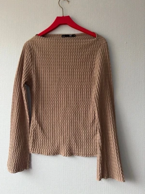 Beige långärmad topp med struktur - Trendig beige topp med lång ärm och vågig struktur i materialet. Modellen har en rak passform och är tillverkad i ett mjukt syntetmaterial. Perfekt att styla med jeans eller kjol för en cool look.